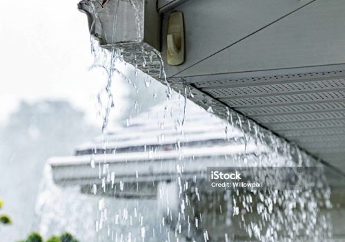 Drenching downpour rain storm water is overflowing off the tile shingle roof - streaming and splashing over the eaves trough aluminum roof gutters on a suburban residential colonial style house near Rochester, New York State, during a torrential mid-summer July downpour.